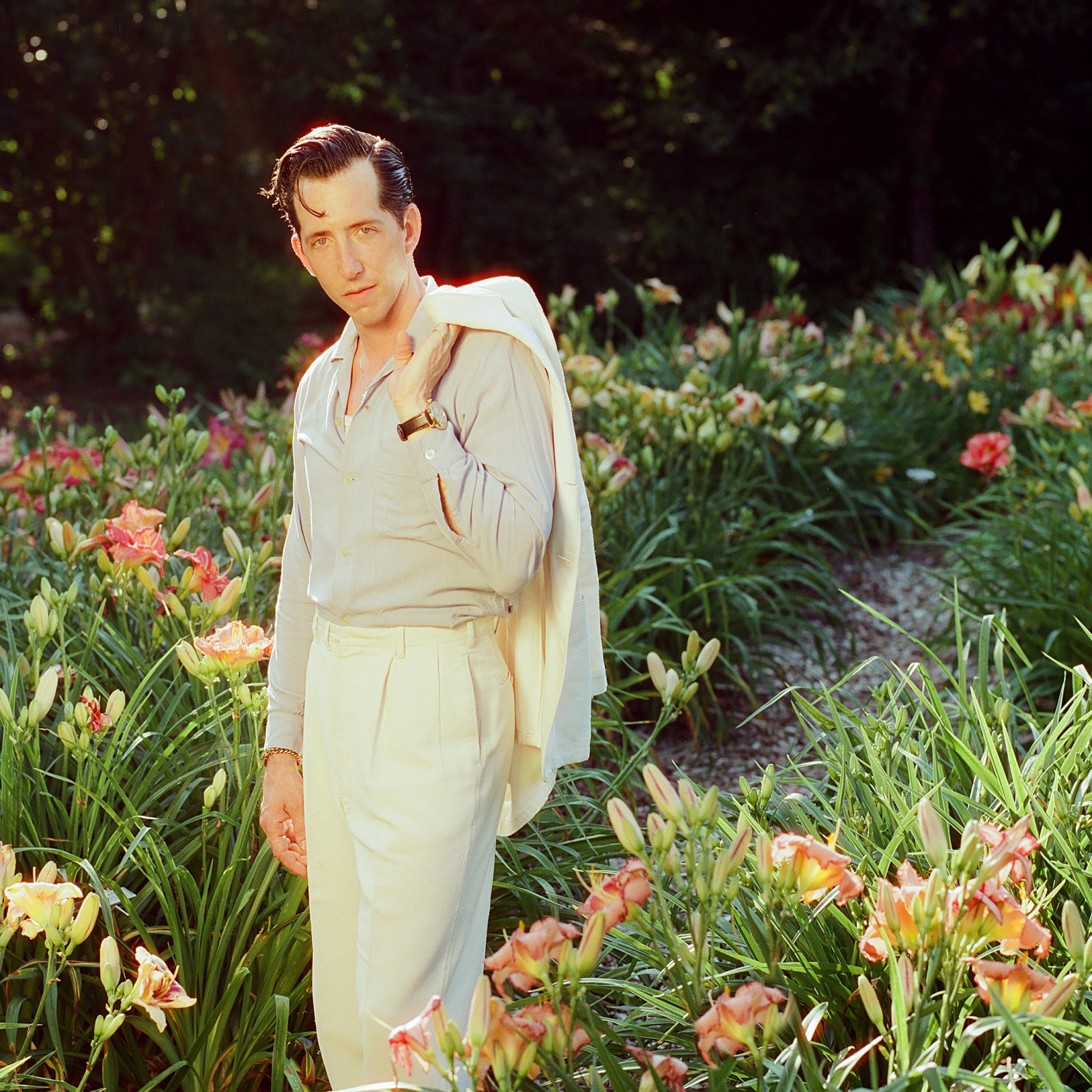 Artist Pokey LaFarge standing in a field of daylilies, wearing cream colored pants holding the collar of the matching suit jacket over his left shoulder, with a light tan button down shirt, open at the neck