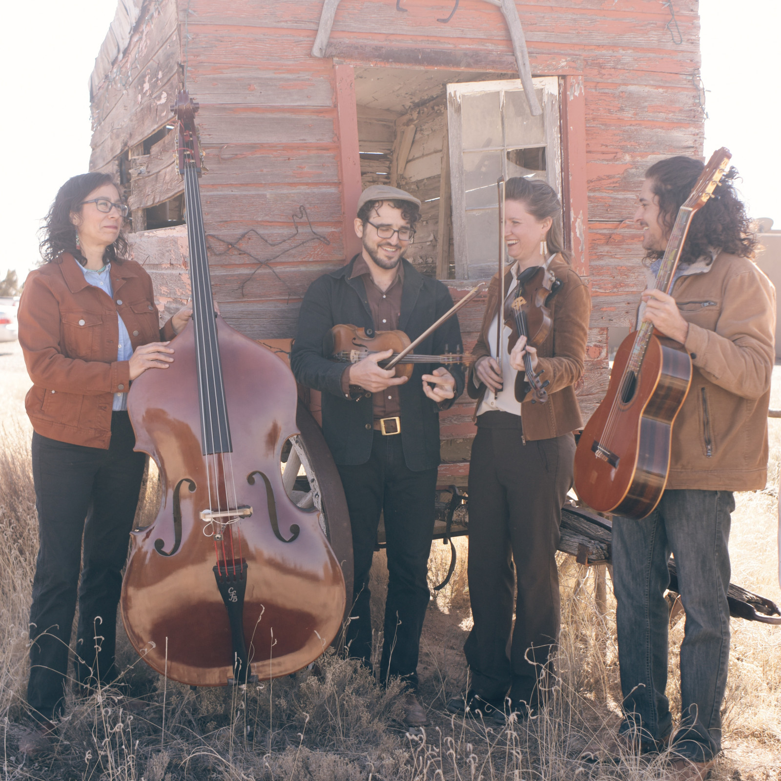 The 4 members of the band Lone Pinon standing in front of an old wooden wagon, holding their instruments: a double bass, two violins and a guitar 