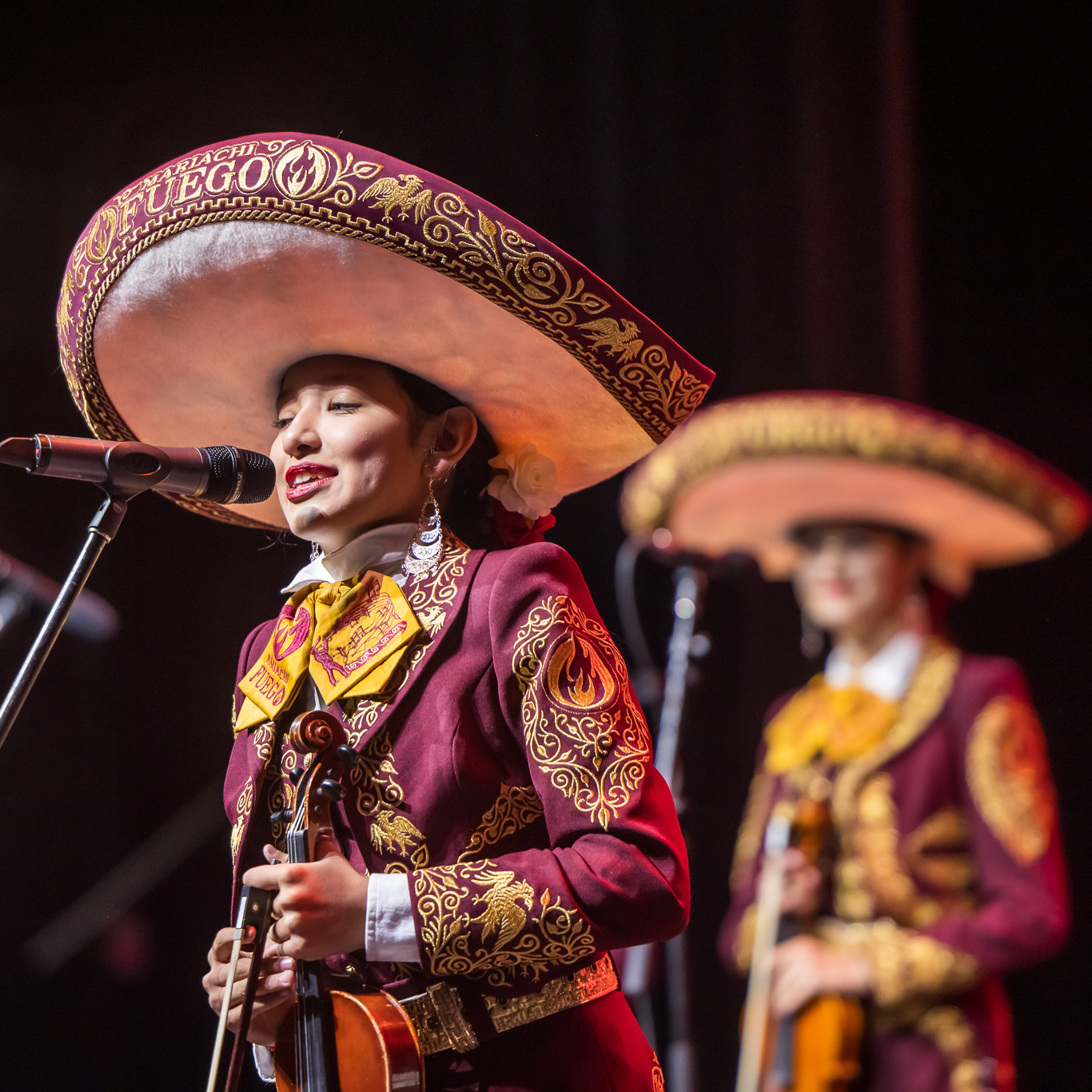 Two members of Mariachi Fuego, holding their violins while one of them is singing or speaking into a microphone